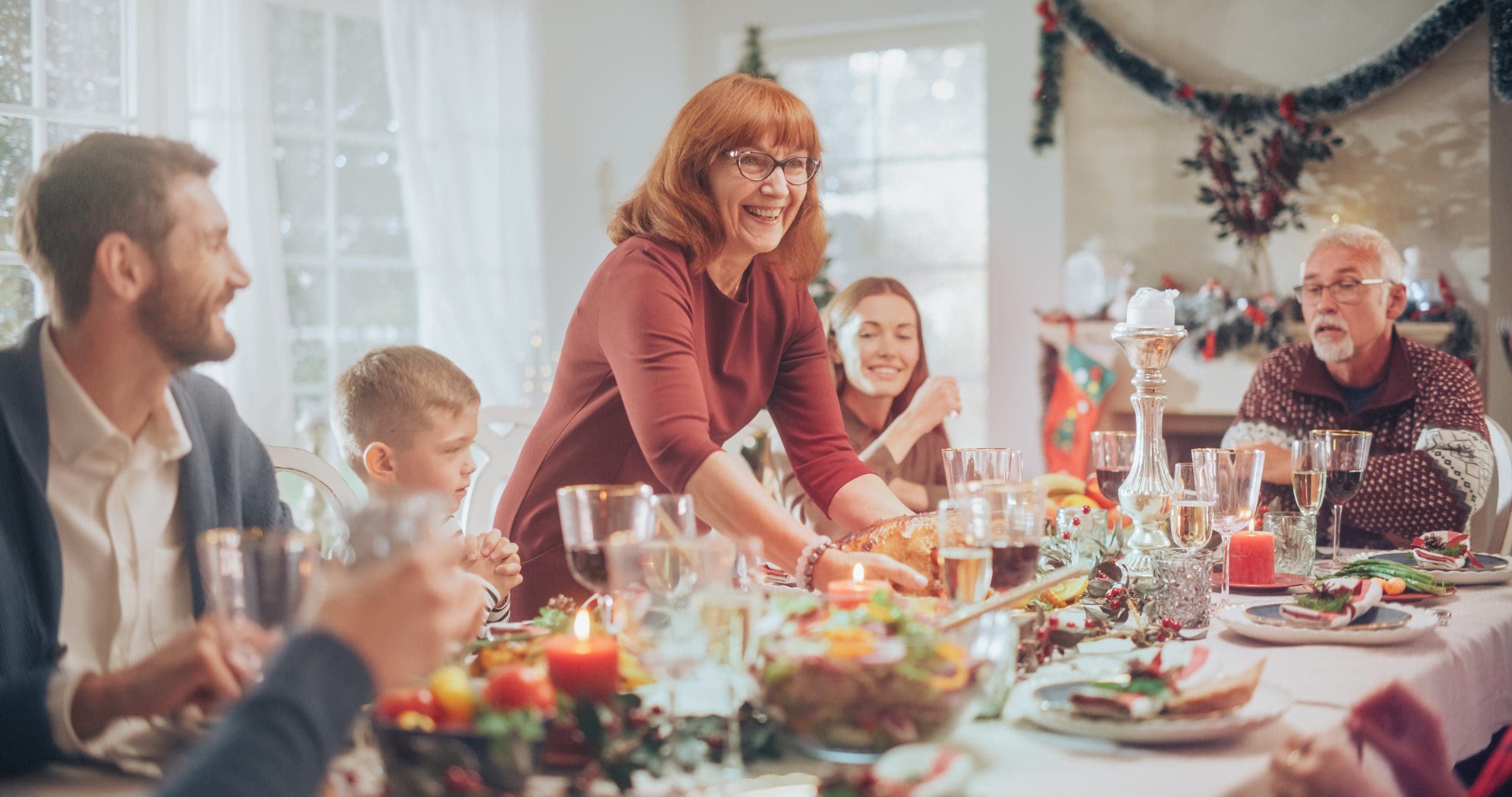 Group of Relatives and Friends Sitting Together Behind a Dining Table with Tasty Meals and Festive Decorations. Senior Woman Bringing a Grilled Chicken, Creating a Joyful Holiday Atmosphere